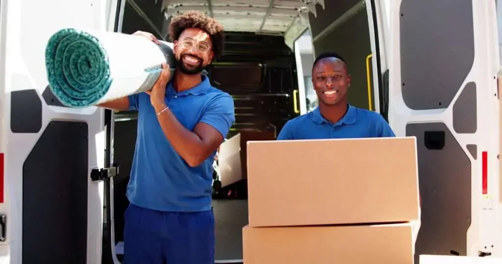 Two professional movers unloading a van, one carrying a rolled rug and the other standing beside stacked moving boxes.