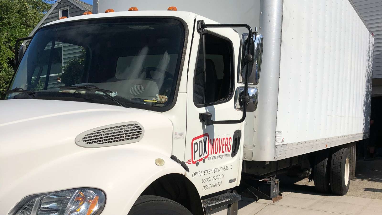 Close-up of a white PDX Movers box truck cab showing the company logo and USDOT licensing information on the driver's side door, parked in a residential driveway.