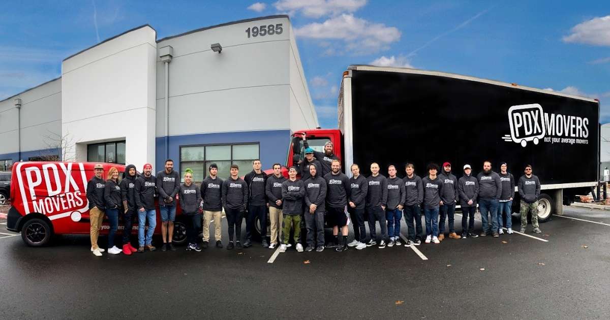 The full PDX Movers team posing together in front of their warehouse, a branded red cargo van, and a large black moving truck with the PDX Movers logo