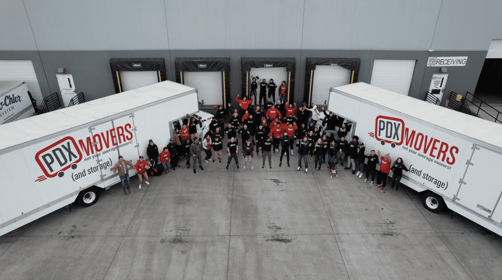 A team photo of employees between two PDX Movers trucks outside a warehouse loading dock.