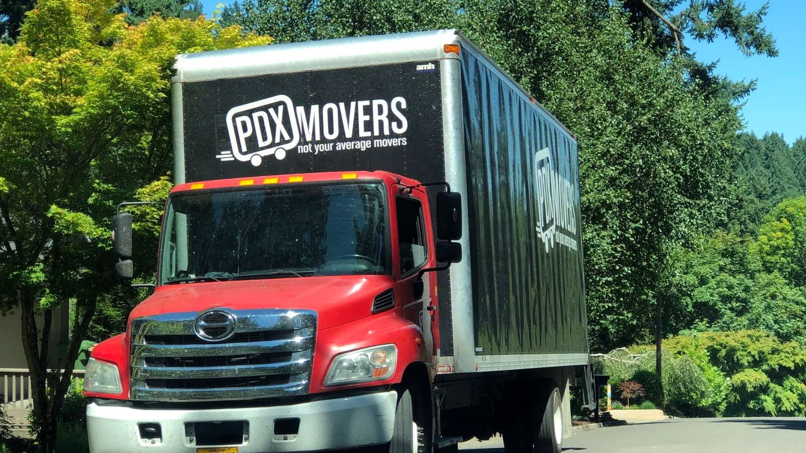 A PDX Movers box truck with a red cab and black branded cargo box displaying the company logo and tagline 'not your average movers,' parked on a tree-lined street on a sunny day.