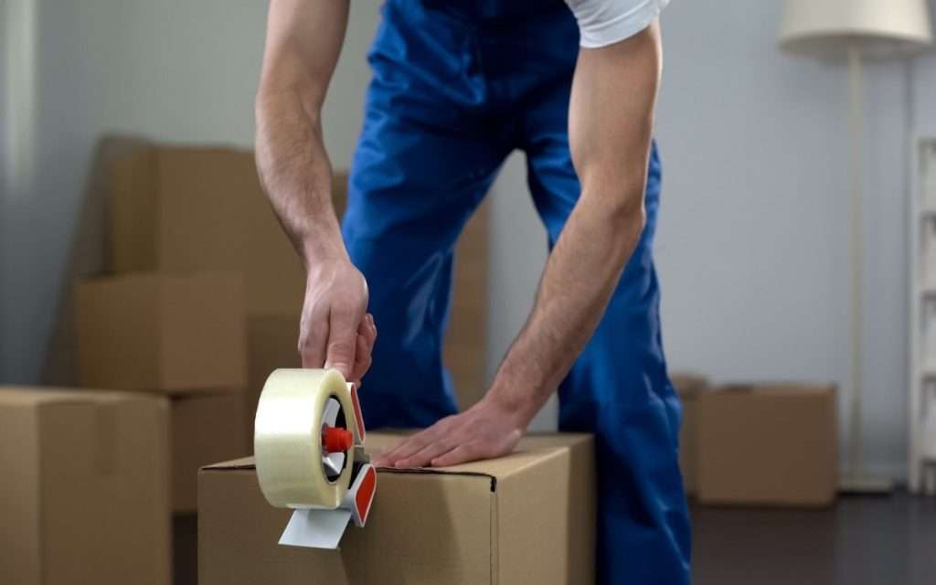 A moving company worker leans over to tape up a box.